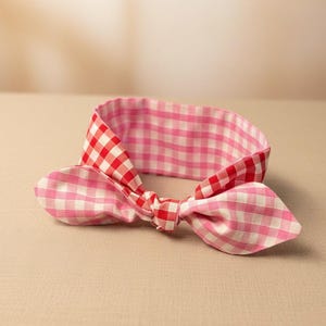 Puede incluir: Una diadema reversible con estampado de cuadros rosas y rojos. La diadema presenta un lazo anudado y un vichy rosa m&aacute;s claro en el reverso. La tela parece ser de algod&oacute;n.