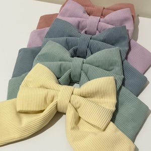 May include: A selection of pastel-coloured corduroy hair bows. The bows are in shades of yellow, green, blue, lavender, and peach. They are arranged in a layered fan shape, highlighting their textured fabric and simple design. Suitable for all ages.