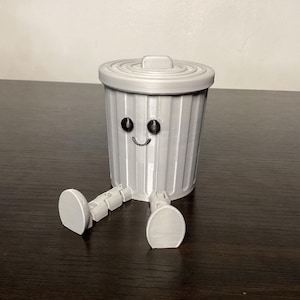 May include: A gray plastic trash can with a smiling face and black eyes. The trash can has legs and is sitting on a dark brown surface.