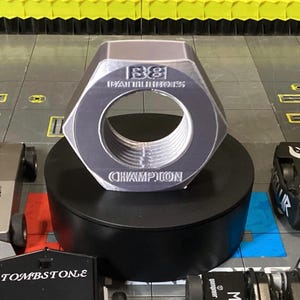 May include: A large, silver, hexagonal nut trophy with the words "BattleBots" and "Champion" inscribed on it. The trophy is sitting on a black pedestal.