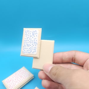 May include: A close-up of miniature beige rectangular food items, resembling pastries. One is held up, showing a white frosting with blue sprinkles. Another, on the bottom, has pink sprinkles. The background is a solid blue.