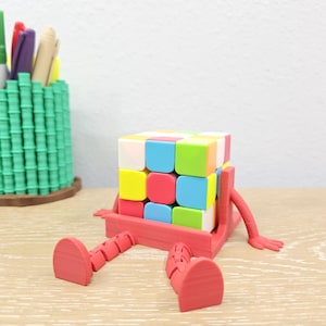 May include: A colorful Rubik's Cube sits in a red holder with arms and legs, giving it a playful appearance. The cube features squares of various colors. A green pen holder with pens is in the background.