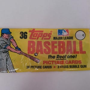 May include: A sealed pack of Topps Major League Baseball cards. The yellow wrapper features a baseball player illustration, the Topps logo, and text that reads "BASEBALL the Real one! PICTURE CARDS". Includes 36 picture cards and 3 sticks of bubble gum.