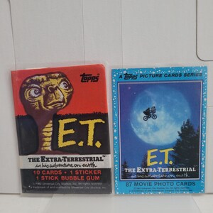 May include: Two vintage E.T. trading card packs. One pack is red with an image of E.T. and the text "E.T. The Extra-Terrestrial in his adventure on earth." The other pack is blue with a scene of E.T. and Elliot flying in front of the moon.