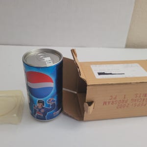 May include: A Pepsi 2002 Racing Program can with a blue, red, and white design, a cardboard box, and a clear plastic container. The can features a race car driver graphic.