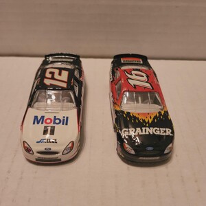 May include: Two toy race cars. One is white with "Mobil 1" and the number "12". The other is black with the number "16" and the word "GRAINGER". Both cars have sponsor logos and are on a light surface.