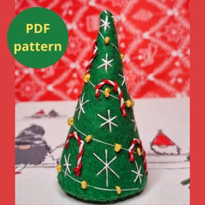 Puede incluir: Un adorno de árbol de Navidad en forma de cono de fieltro verde con copos de nieve blancos, bastones de caramelo rojos y blancos y luces amarillas. El adorno está sobre una superficie blanca con un fondo rojo y blanco.