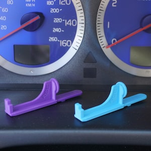 May include: Two plastic phone holders, one purple and one blue, are sitting on a black dashboard. The dashboard has two gauges, one with a red needle pointing to 40 and the other with a red needle pointing to 0.