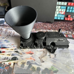 May include: A black plastic funnel attached to a black engine component. The engine component has the word "VOLVO" and the numbers "6420419" and "1125366" printed on it. The funnel is cone-shaped.