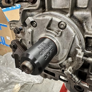 Op de afbeelding: Close-up van een motoronderdeel met een zwart cilindrisch gereedschap met de tekst "RS CRANK DRIFT" dat in het motorblok is geschroefd. De motor is metaalgrijs met diverse bouten en mechanische onderdelen.