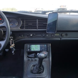Può includere: Vista interna del cruscotto di un'auto d'epoca. Il cruscotto nero presenta indicatori, bocchette dell'aria e una radio con schermo di navigazione. Uno smartphone è montato sul cruscotto. Sono visibili anche il volante e la leva del cambio.