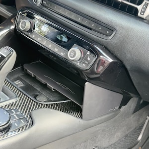 Puede incluir: Interior de coche negro con consola central que incluye radio, controles de aire acondicionado y una bandeja de almacenamiento. La consola tiene un acabado brillante y está rodeada de molduras de fibra de carbono.