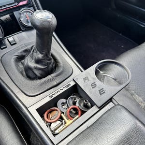 May include: A black car interior with a black leather gear shift and a black plastic storage compartment with the word "Porsche" printed on it. The compartment is filled with various small objects, including screws, washers, and o-rings. The compartment also has a cup holder with the letters "RSE" printed on it.