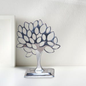 May include: A silver metal tree sculpture with a cutout design. The tree has a wide base and a tall, slender trunk with many branches.
