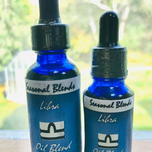 May include: Two cobalt blue glass bottles with black droppers. The bottles have labels that read "Seasonal Blends Libra Oil Blend". One bottle is labeled "One Fluid Ounce" and the other "One-Half Fluid Ounce".