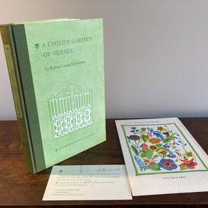 May include: A green hardcover book titled "A Child's Garden of Verses" by Robert Louis Stevenson. The book features a white illustration of a gate on the front cover. The book is sitting on a wooden surface with a white card that says "A Very Special Edition" and a smaller white card with text about the book.