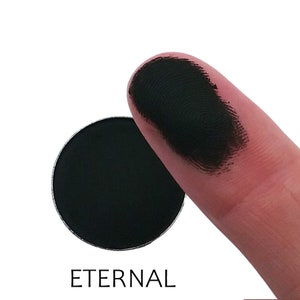 May include: A black eyeshadow pan with the word "ETERNAL" printed below. A fingertip shows a swatch of the black eyeshadow.