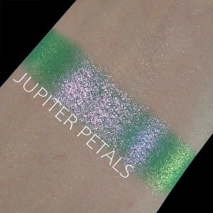 May include: A close-up of a green and silver eyeshadow swatch on skin. The swatch is labeled "Jupiter Petals".