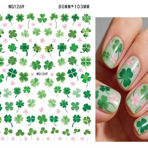 May include: Nail art stickers featuring green shamrocks and pink flowers. The sheet measures 80mm x 103mm. The image also shows a hand with nails decorated with shamrocks and flowers.