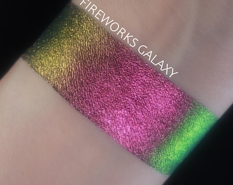 Può includere: Un campione di ombretto iridescente in un gradiente di colori, tra cui giallo, rosa e verde. Il testo "FIREWORKS GALAXY" è stampato in bianco sulla parte superiore del campione.