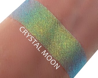 May include: A close-up of a green, blue, and yellow iridescent eyeshadow swatch on a person's arm. The text "CRYSTAL MOON" is written below the swatch.