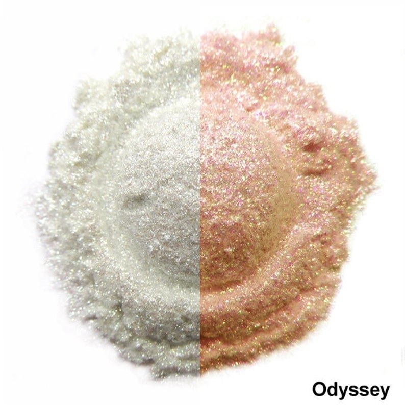 Pu&ograve; includere: Primo piano di un mucchio di glitter iridescente, diviso a met&agrave;. Un lato &egrave; bianco, l'altro &egrave; rosa pallido con un effetto perlescente. Il testo "Odyssey" &egrave; stampato sotto il glitter.