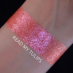 Puede incluir: Una muestra de maquillaje brillante y con purpurina en tonos coral y rosa, con el texto "READ MY TULIPS" en blanco. El maquillaje se aplica sobre la piel, sobre un fondo oscuro.