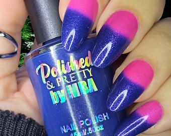 May include: A bottle of blue nail polish with the text "Polished & Pretty by MBA" is held near a hand with nails painted in a pink and blue gradient. The nail polish bottle is 15 ml / .50 oz.