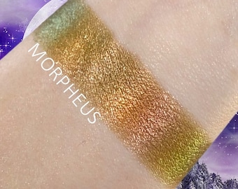 May include: A close-up of a shimmery green and gold eyeshadow swatch on a person's arm. The eyeshadow has a metallic finish and is labeled "MORPHEUS".