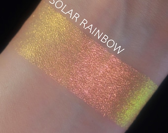 Può includere: Un campione di trucco scintillante con un effetto sfumato, che passa dall'oro al pesca e termina con una tonalità verde lime. Il testo "SOLAR RAINBOW" è stampato in bianco sulla parte superiore del campione.