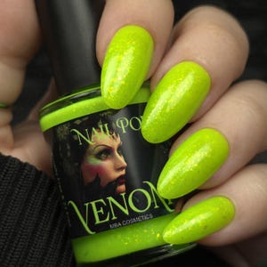 May include: A bottle of "Venom" nail polish by MBA Cosmetics, held next to painted nails. The polish is a bright neon green with gold glitter. The bottle label features a woman's face with green and red accents.