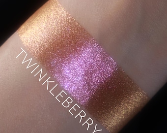May include: A swatch of shimmery eyeshadow with a pink and gold color shift. The text "TWINKLEBERRY" is written on the swatch.