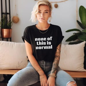 May include: Black crew neck t-shirt with the white text "none of this is normal." The person is wearing blue jeans with ripped knees and has multiple bracelets on their wrist. The shirt is a casual style.