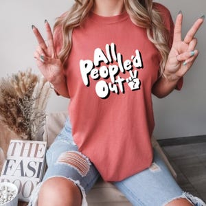 May include: A coral-coloured t-shirt featuring the text "All People'd Out" in white with a black outline and a peace sign. The shirt is worn with light-wash denim jeans.