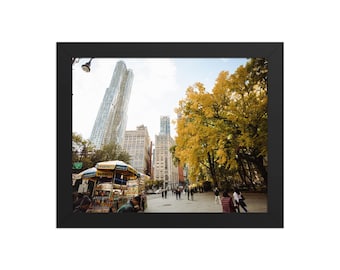 May include: A framed photograph depicting a city street with tall buildings and people. A food cart is visible, and the trees have yellow leaves, indicating autumn. The sky is overcast.
