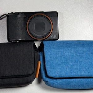 May include: A black digital camera with a brown lens ring and the text "GR" sits above two camera cases. One case is black, and the other is blue. Both cases have a flap closure and a brown leather accent.