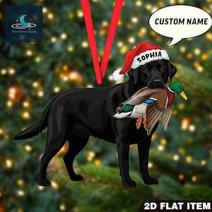 May include: A black Labrador Retriever ornament wearing a Santa hat with the name "Sophia" on it, holding a duck in its mouth. The ornament has a red ribbon for hanging and the text "CUSTOM NAME" in a speech bubble.