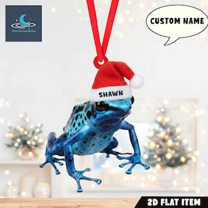 May include: A blue frog ornament with a Santa hat that says "Shawn". The ornament is hanging from a red ribbon. The ornament is a 2D flat item. The image also includes the text "Custom Name".