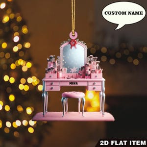 May include: A pink vanity ornament with a mirror, drawers, and a stool. The vanity is adorned with makeup brushes and products. The name "MINA" is displayed on the front. A speech bubble above says "CUSTOM NAME". The ornament is suspended by a gold chain.
