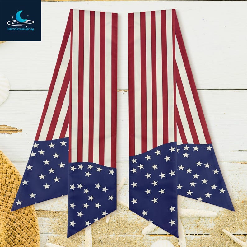 Pu&ograve; includere: Due sciarpe con un design ispirato alla bandiera americana. Ogni sciarpa presenta una sezione blu con stelle bianche e strisce rosse e bianche. Sono esposte su una superficie di legno bianco con sabbia e conchiglie.