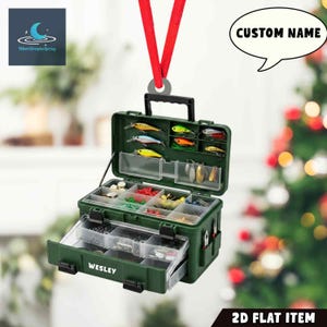 May include: A green fishing tackle box ornament with the name "WESLEY" on the drawer. The box is open, revealing various fishing lures and compartments. A red ribbon is attached for hanging. Text on the image says "CUSTOM NAME" and "2D FLAT ITEM".