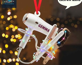 May include: A white and rose gold hair dryer, flat iron, and brush ornament with the name "GEMMA" printed on the hair dryer. The ornament is decorated with a string of colorful lights and a red ribbon for hanging. The text "CUSTOM NAME" is in a speech bubble.