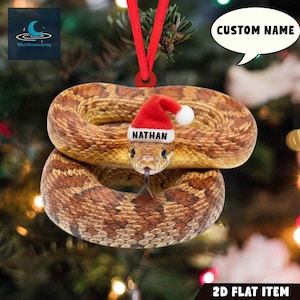 May include: A 2D flat Christmas ornament featuring a coiled snake with a red Santa hat. The hat has the name "NATHAN" printed on it. The ornament has a red ribbon for hanging and the words "CUSTOM NAME" in a speech bubble.