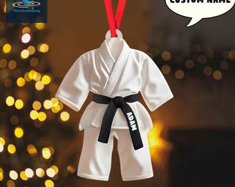 May include: A white karate uniform ornament with a black belt and the name "ADAM" printed on it. The ornament is hanging from a red ribbon. A speech bubble says "CUSTOM NAME".