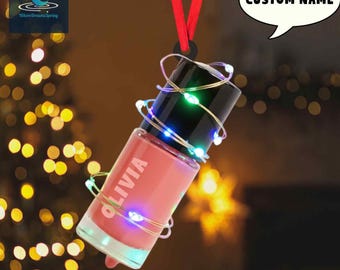 May include: A pink nail polish bottle ornament with the name "OLIVIA" printed on the side. The bottle is wrapped with a string of colorful LED lights and suspended by a red ribbon. A speech bubble says "CUSTOM NAME".
