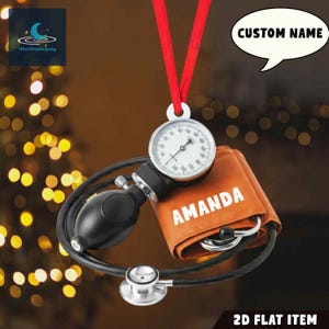 May include: A medical-themed ornament featuring a blood pressure gauge, cuff with the name "AMANDA", and stethoscope. The ornament hangs from a red ribbon. The gauge displays numbers and a needle. The background is blurred with bokeh lights.