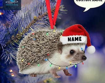 May include: A Christmas ornament featuring a hedgehog wearing a Santa hat with the word "NAME" on it. The hedgehog is decorated with a string of colorful lights and has a red ribbon for hanging. The text "CUSTOM NAME" is in a speech bubble.