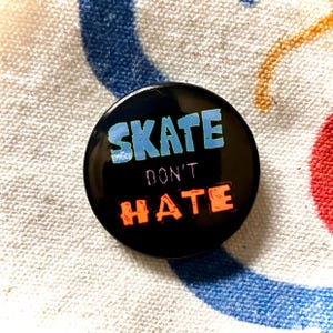 May include: A black button with the words "SKATE DON'T HATE" in blue and orange lettering. The word "SPRUCE" is printed in small green letters above the word "SKATE". The button is on a white and blue striped surface.