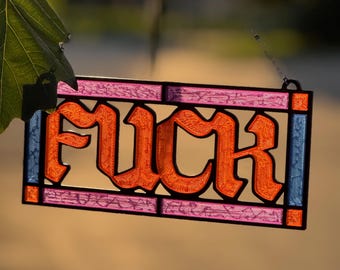 May include: A rectangular stained glass suncatcher featuring the word FUCK in bold orange lettering. The piece is framed in black metal with pink textured glass panels above and below the text, and blue textured glass panels on the sides.