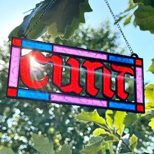 May include: A rectangular stained glass suncatcher featuring the word "cunt" in bold red lettering. The piece is framed by a black metal border with blue and purple glass accents and is suspended by a thin black metal chain for hanging.